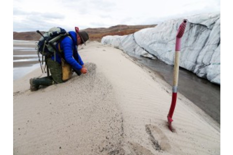 Research Kurt Kjær kneels on all fours on a ridge of sand in front of the glacier's edge.