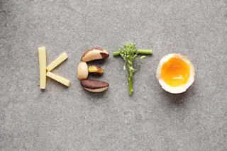 The word "KETO" spelled with food items on a grey background: "K" with fries, "E" with Brazil nuts, "T" with broccoli, and "O" with a half-boiled egg