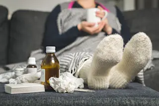  sick woman drinking hot beverage to ease flu symptoms, fever and virus. Dirty paper towels, cough medicine, and tissues on table. Ill person 