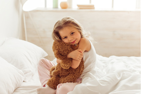 Sweet little girl is hugging a teddy bear, looking at camera and smiling while sitting on her bed at home