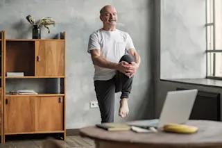 Senior sportman doing stretching and balancing yoga exercise at home using online lesson on notebook.
