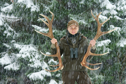 woman holding shed antlers in front of an evergreen in the snow
