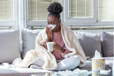 woman with blanket on couch blowing nose and holding tea