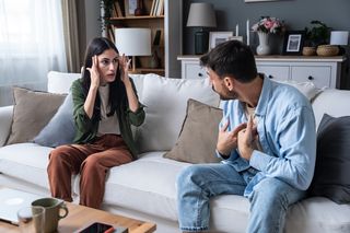 young couple sitting on the couch. The woman has her hands on her temples and the man is pointing at his chest. 