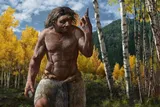 An ancient human stands in a forest, holding a stone and a stone point. 