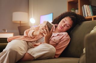 a young woman sits on her couch scrolling on her phone 