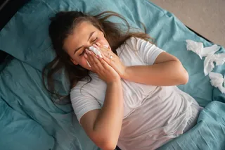 A woman laying in bed and blowing her nose surrounded by tissues A woman laying in bed and blowing her nose surrounded by tissues