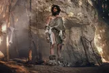 An early human man wearing furs and standing in a cave near wooden tools