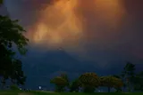 A wildfire burns across a forested hillside beneath a towering orange-brown cloud of smoke.