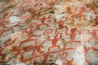 Red Huashan rock paintings of human figures, animals, and circular symbols cover a weathered limestone cliff in southern China.