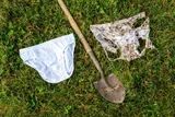 A clean white pair of cotton underwear next to a garden shovel and a badly decomposed, soil-stained pair after two months buried underground